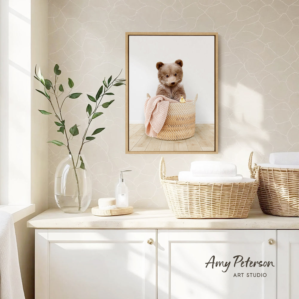 a framed photograph of a bear sitting in a woven basket, placed on a wall above a white cabinet. The cabinet has two baskets on it, and there is a vase with green leaves on the countertop.
