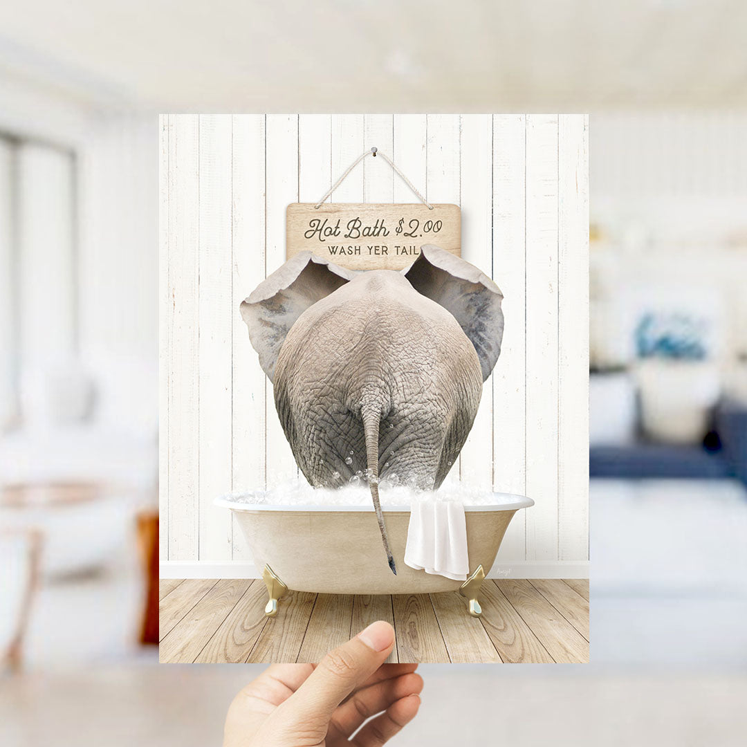 A hand holding a framed image of an elephant sitting in a bathtub, with a sign above it that says "Hot Bath $2.00 WASH YOUR TAIL".