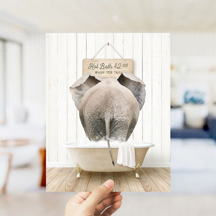 A hand holding a framed image of an elephant sitting in a bathtub, with a sign above it that says "Hot Bath $2.00 WASH YOUR TAIL".