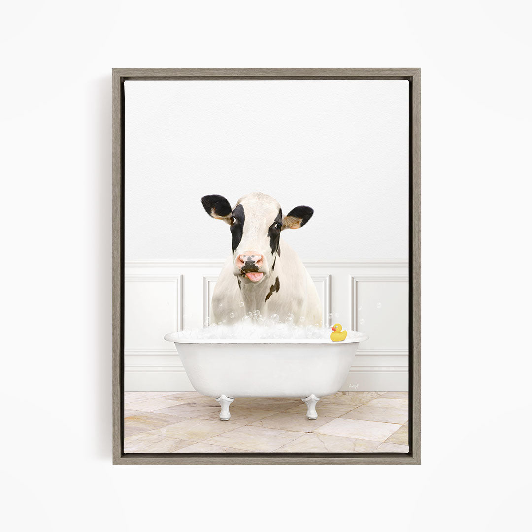 A black and white cow is sitting in a white bathtub filled with bubbles, with a rubber duck floating in the water.