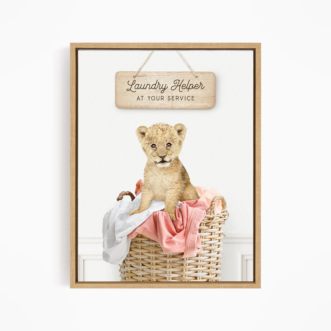 A young lion cub sitting in a wicker basket filled with clothes, with a wooden sign above that reads "Laundry Helper at your service".