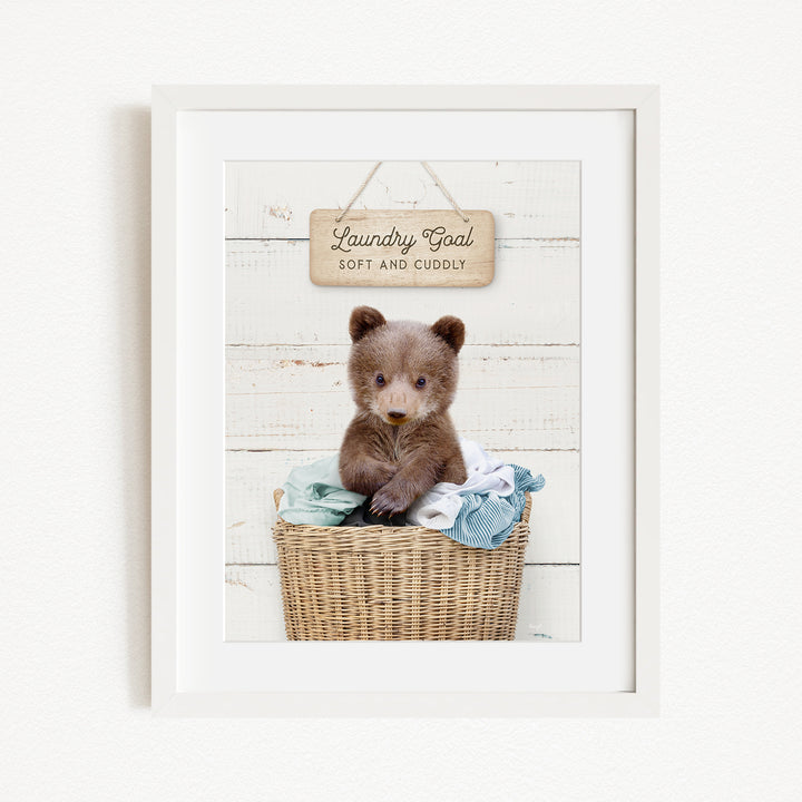 A cute brown bear sitting in a wicker laundry basket, with a wooden sign above that says "Laundry Goal Soft and Cuddly".
