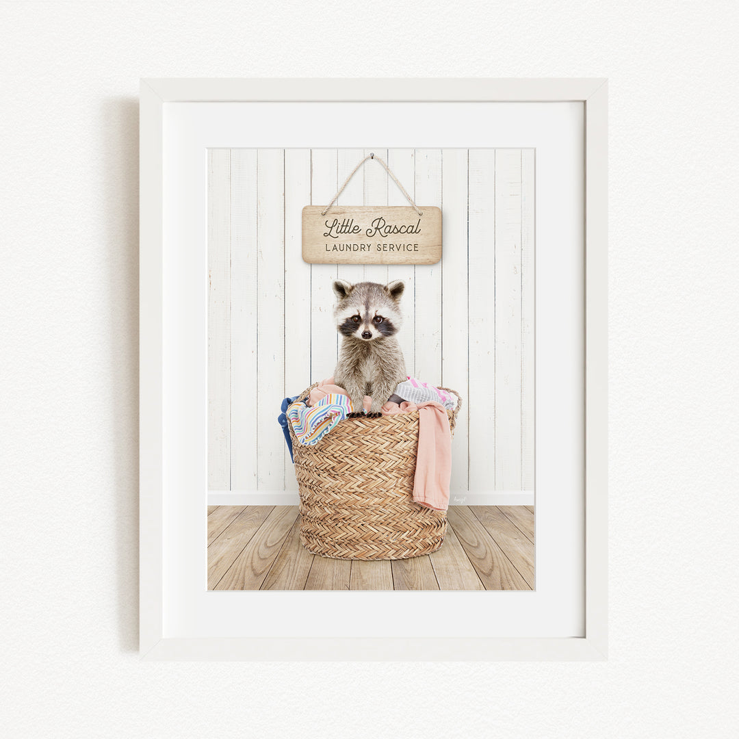 A raccoon sitting in a woven laundry basket, with a sign above that reads "Little Racquel Laundry Service".