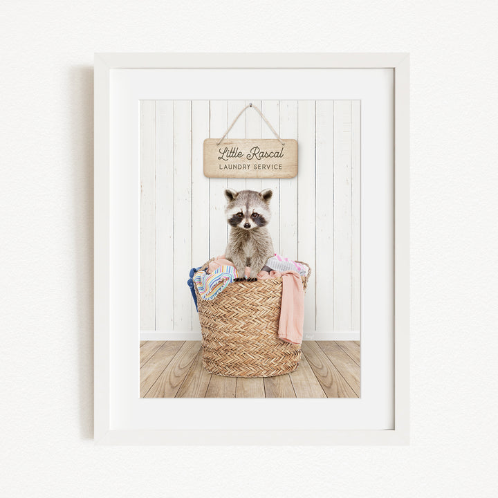 A raccoon sitting in a woven laundry basket, with a sign above that reads "Little Racquel Laundry Service".