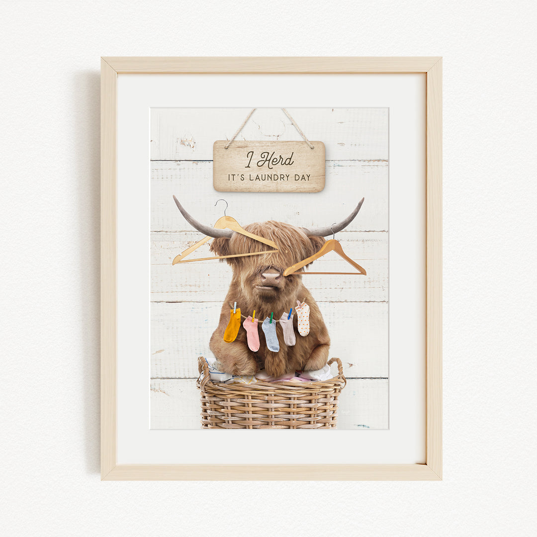 A framed image depicting a Highland cow sitting in a wicker basket, surrounded by clothes and a sign that reads "It's laundry day".