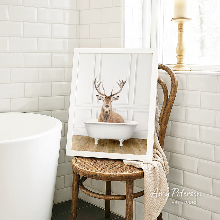 A framed image of a deer sitting in a bathtub is displayed on a wooden chair in a bathroom setting.