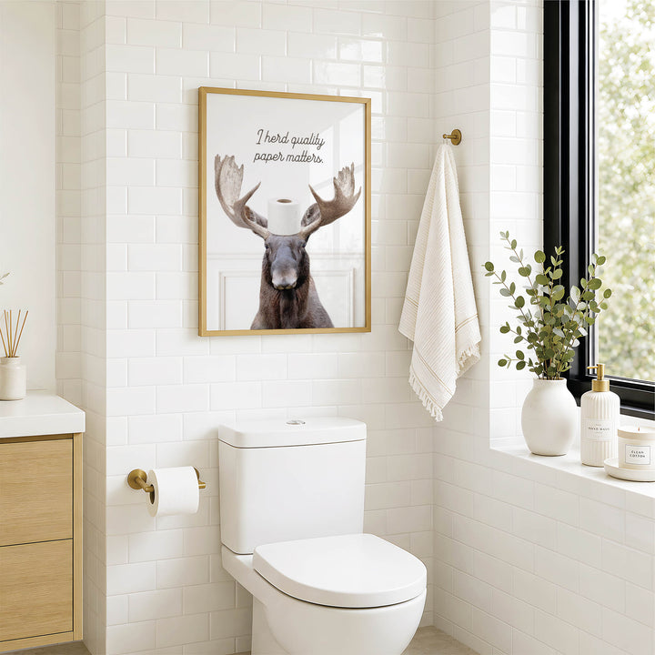 A bathroom with a white toilet, a framed picture of a moose with a toilet paper holder, and a towel hanging on a rack.
