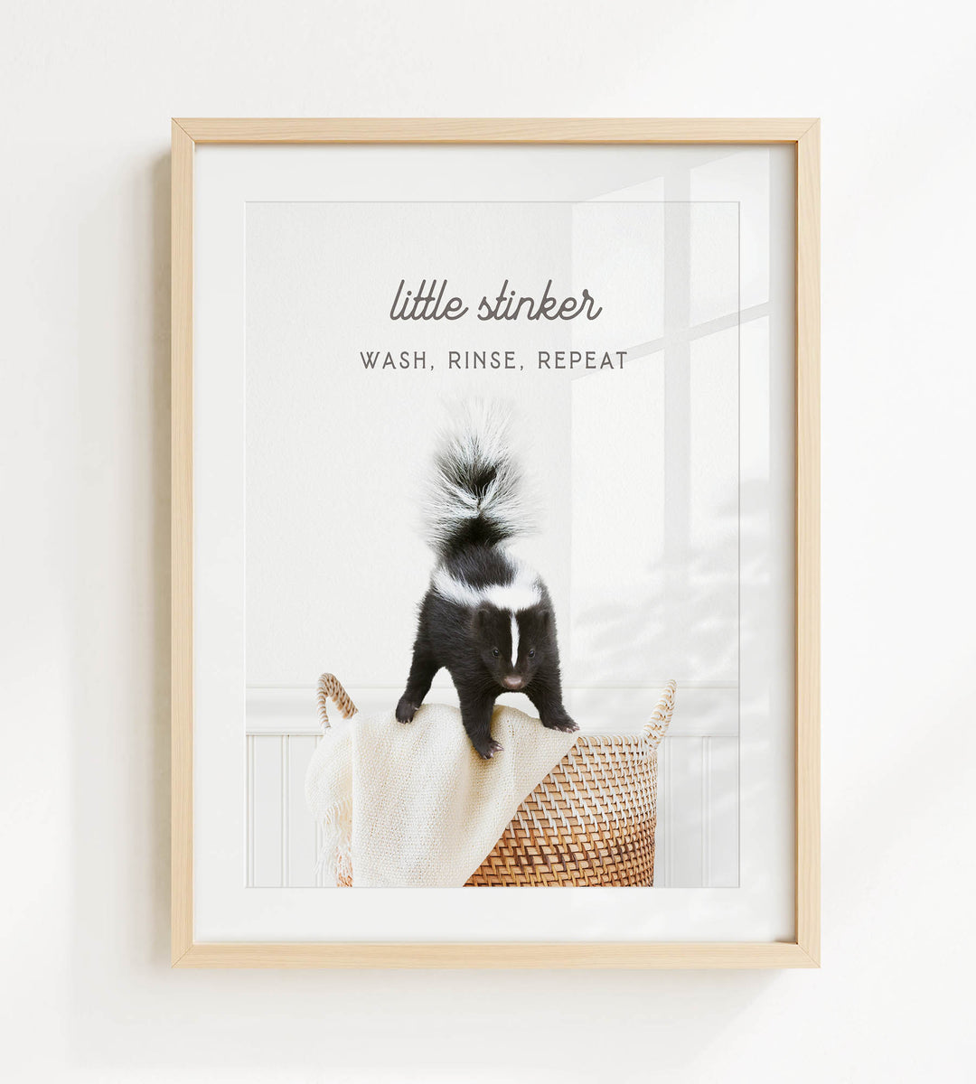 A framed image of a skunk sitting on a woven basket, with the text "Little stinker" and "WASH, RINSE, repeating" written above it.