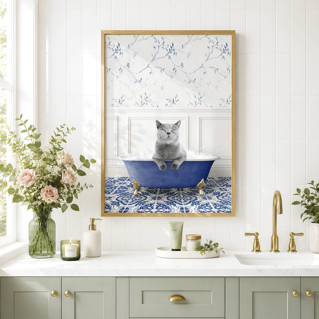 A framed image of a cat sitting in a blue bathtub, surrounded by flowers and a decorative vase, is displayed on a white tiled wall above a bathroom counter with various toiletries and a plant.