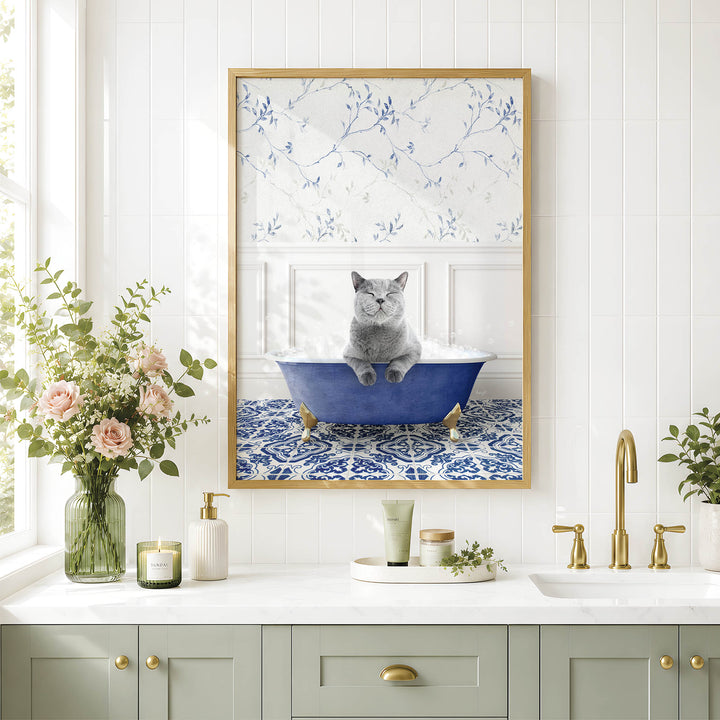 A framed image of a cat sitting in a blue bathtub, surrounded by flowers and a decorative vase, is displayed on a white tiled wall above a bathroom counter with various toiletries and a plant.