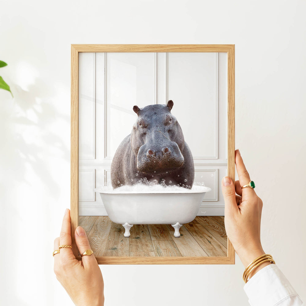 A person's hands are holding a framed image of a hippopotamus sitting in a bathtub filled with water, creating a whimsical and surreal scene.