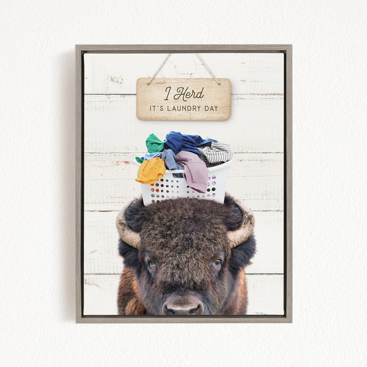 A framed image of a bison with a basket of laundry on its head, accompanied by a wooden sign that says "I herd it's laundry day".