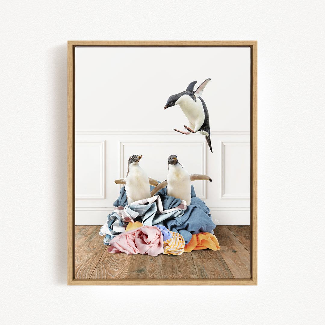 A framed photograph depicting two penguins standing on a pile of clothes, with one penguin flying in the air above the pile.