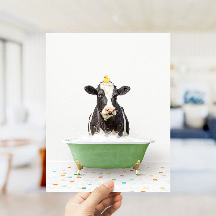 A black and white cow is sitting in a green bathtub filled with bubbles, with a yellow duck perched on its head.