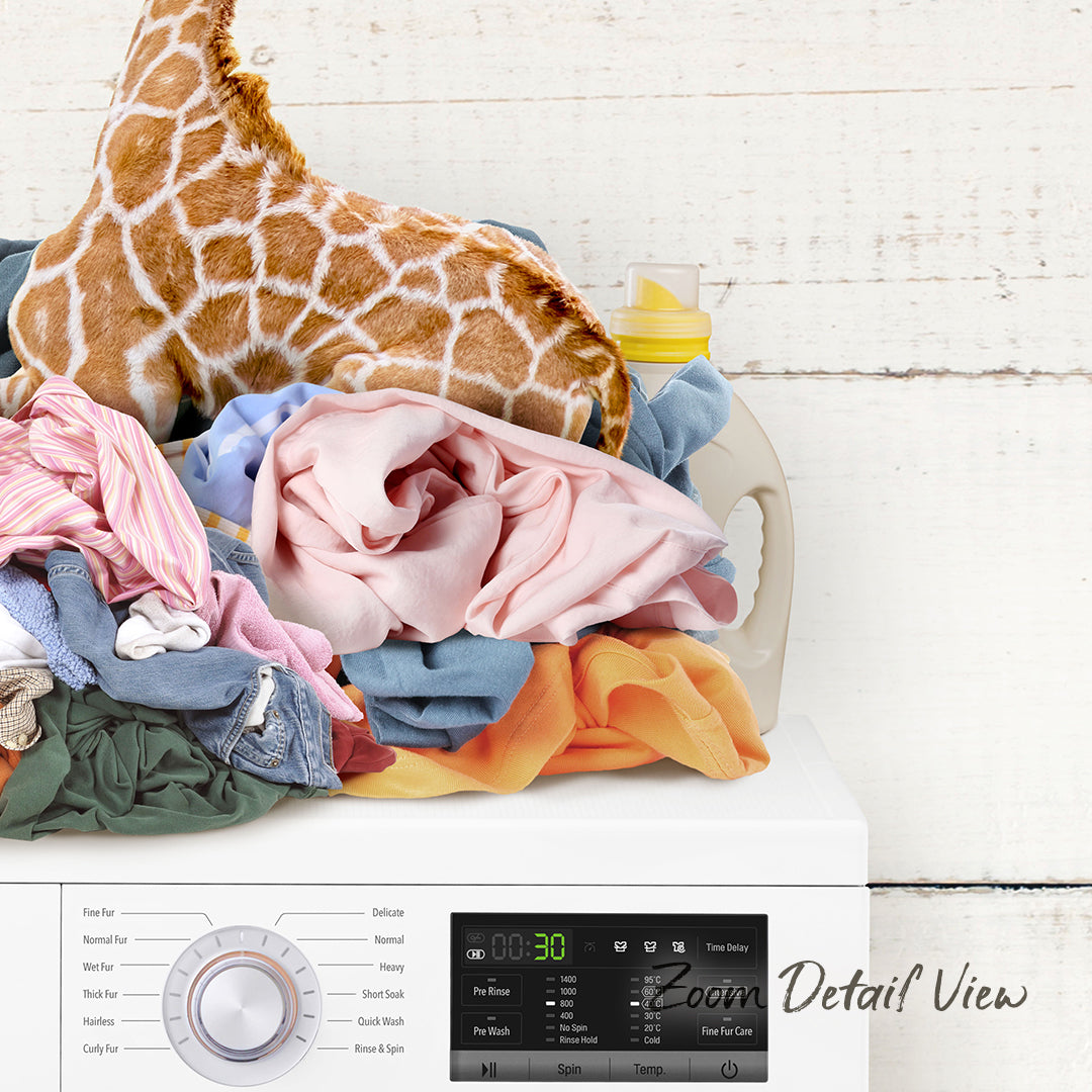 A pile of laundry, including clothes and a stuffed giraffe, sits atop a white washing machine with various settings visible.