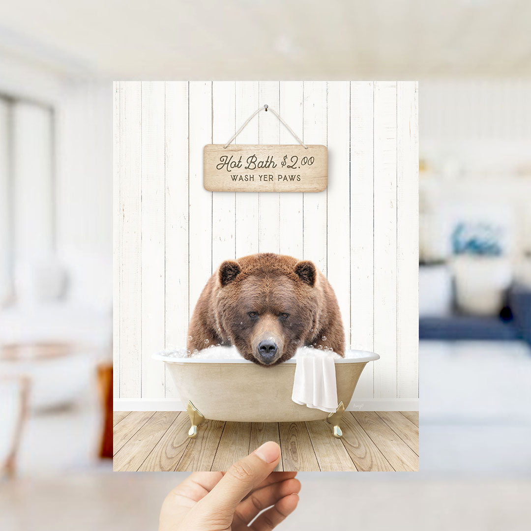 A hand holds up a framed image of a brown bear sitting in a bathtub, with a sign above it that reads "Hot Bath $2.00 WASH YOUR Paws".