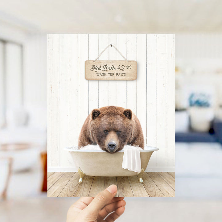 A hand holds up a framed image of a brown bear sitting in a bathtub, with a sign above it that reads "Hot Bath $2.00 WASH YOUR Paws".