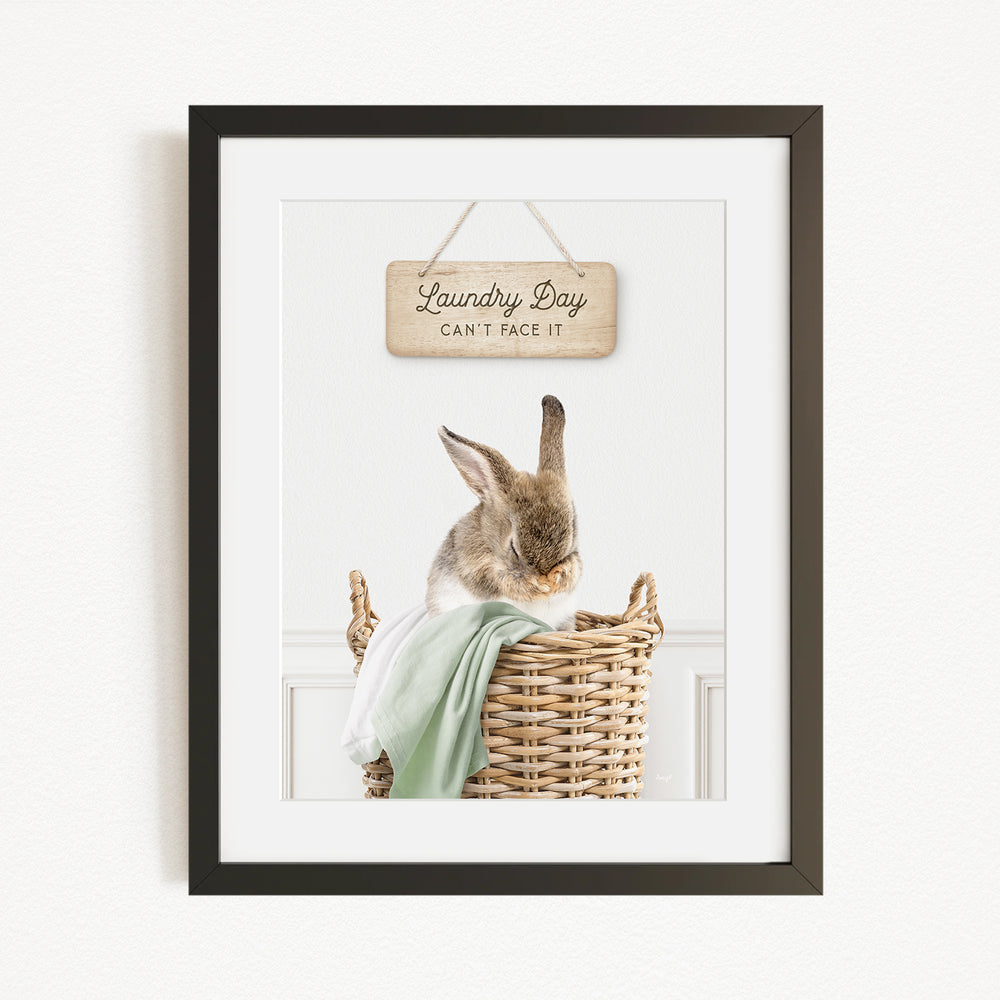A framed image of a rabbit sitting in a wicker basket, with a wooden sign above it that says "Laundry Day Can't Face It".