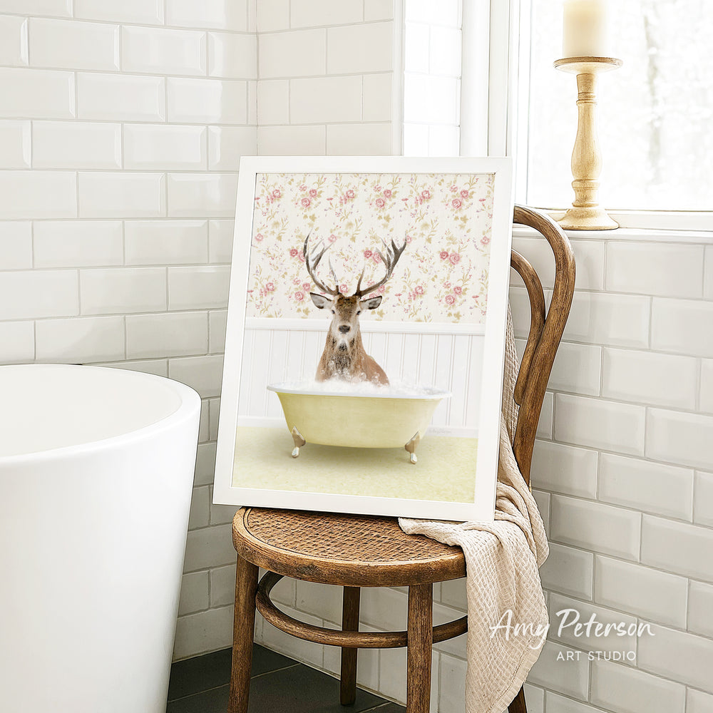 A framed image of a deer with antlers sitting in a bathtub, surrounded by a floral pattern, is displayed on a wooden chair in a bathroom setting.