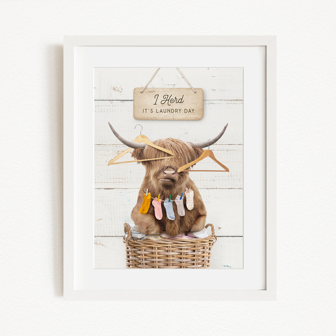 A framed image depicting a cow with horns, surrounded by clothes and hanging clothes, with a sign above that reads "I herd it's laundry day".
