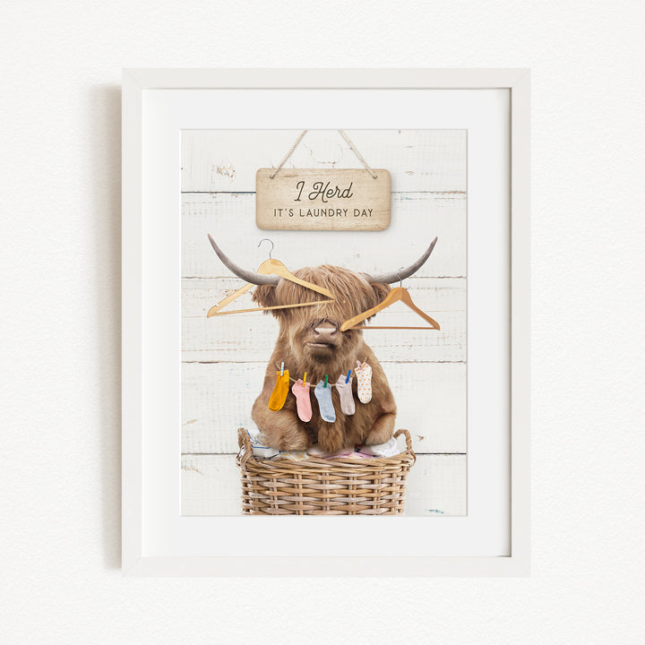 A framed image depicting a cow with horns, surrounded by clothes and hanging clothes, with a sign above that reads "I herd it's laundry day".