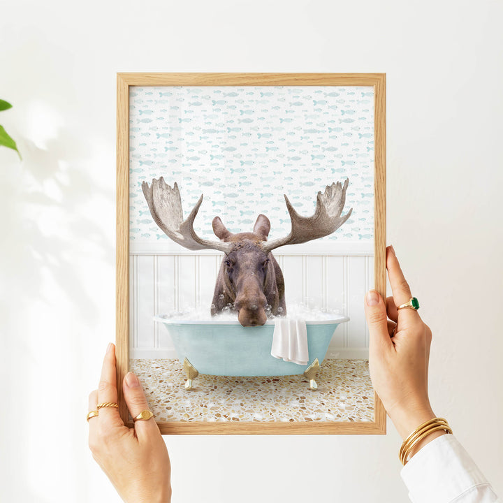 A person's hands are holding a framed image of a moose sitting in a bathtub filled with bubbles, surrounded by a pattern of fish.