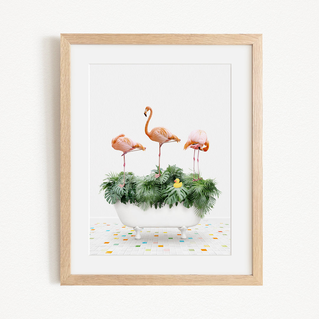 A framed image depicting three flamingos standing on a white bathtub filled with green plants and flowers, set against a white background.