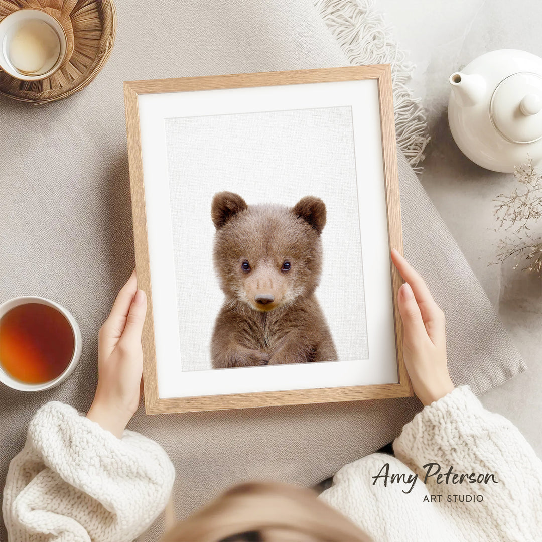 A person is holding a framed picture of a cute brown bear, with a teapot, a cup of tea, and a bowl of tea visible in the background.
