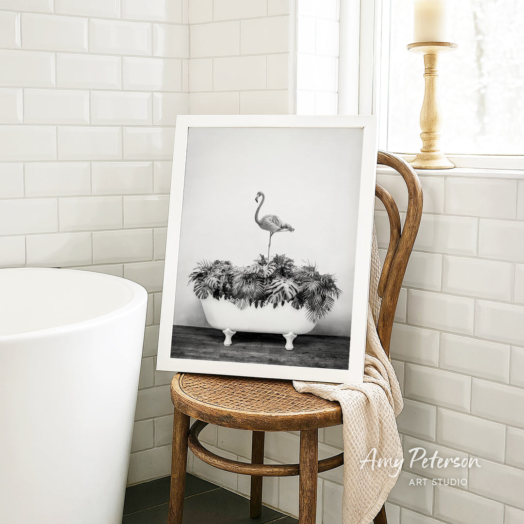 A framed black and white photograph of a flamingo standing in a bathtub filled with plants is displayed on a wooden chair in a bathroom setting.