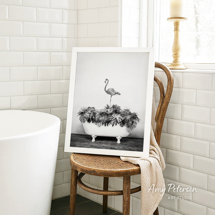A framed black and white photograph of a flamingo standing in a bathtub filled with plants is displayed on a wooden chair in a bathroom setting.