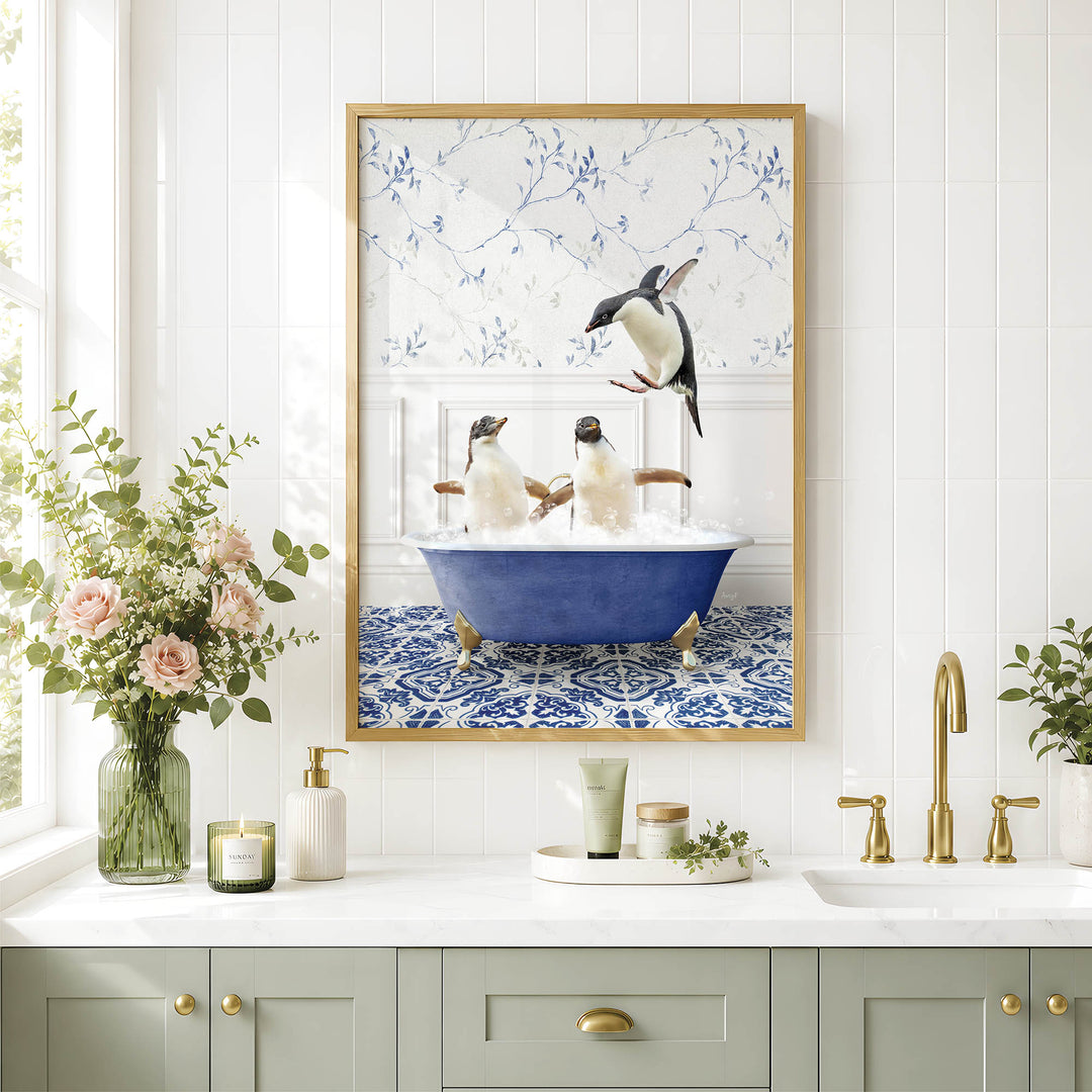 A framed image of two penguins sitting in a blue bathtub, surrounded by flowers, is displayed on a white wall above a bathroom counter with various toiletries and a vase of flowers.