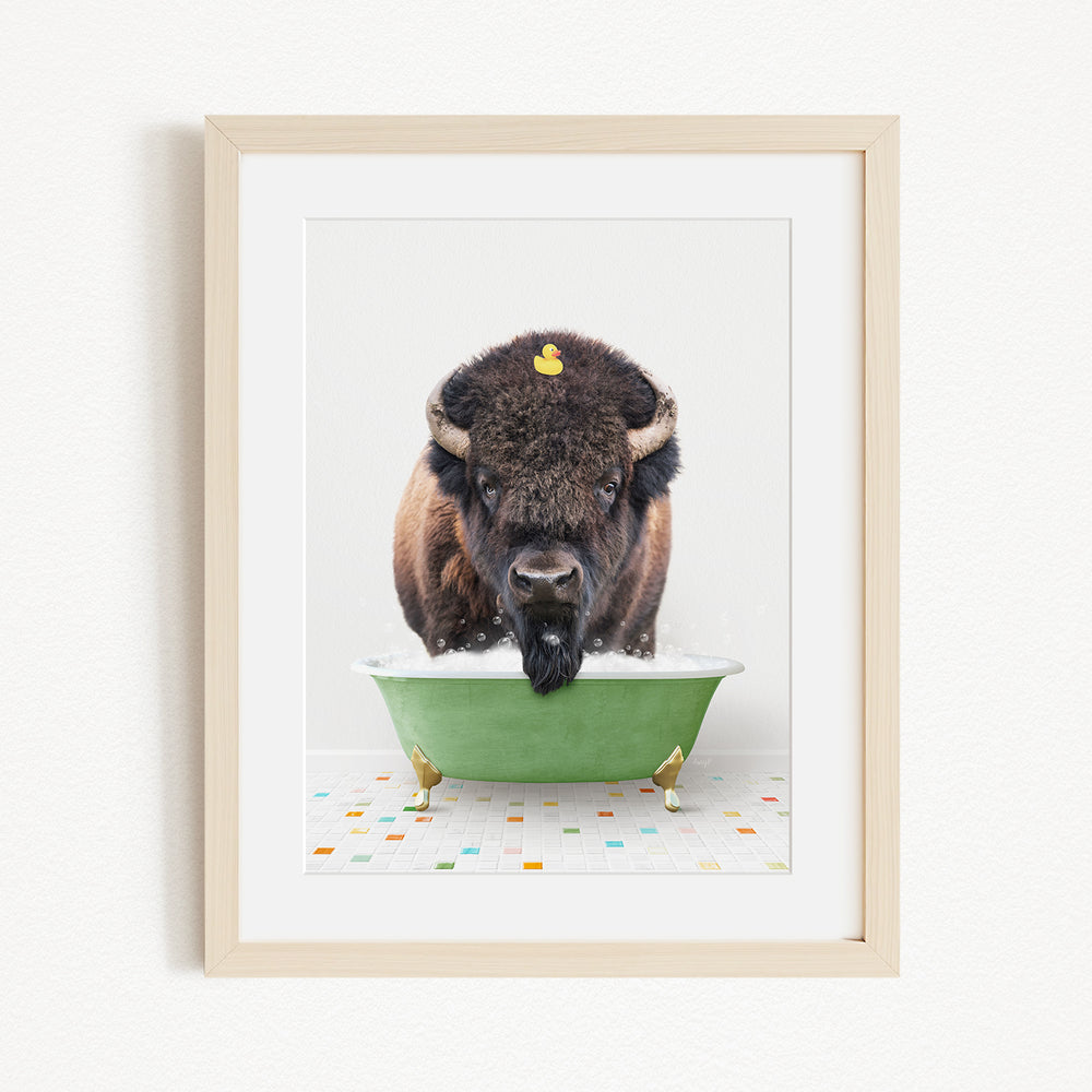 A buffalo with a yellow rubber duck perched on its head is sitting in a green bathtub, surrounded by colorful tiles on the floor.