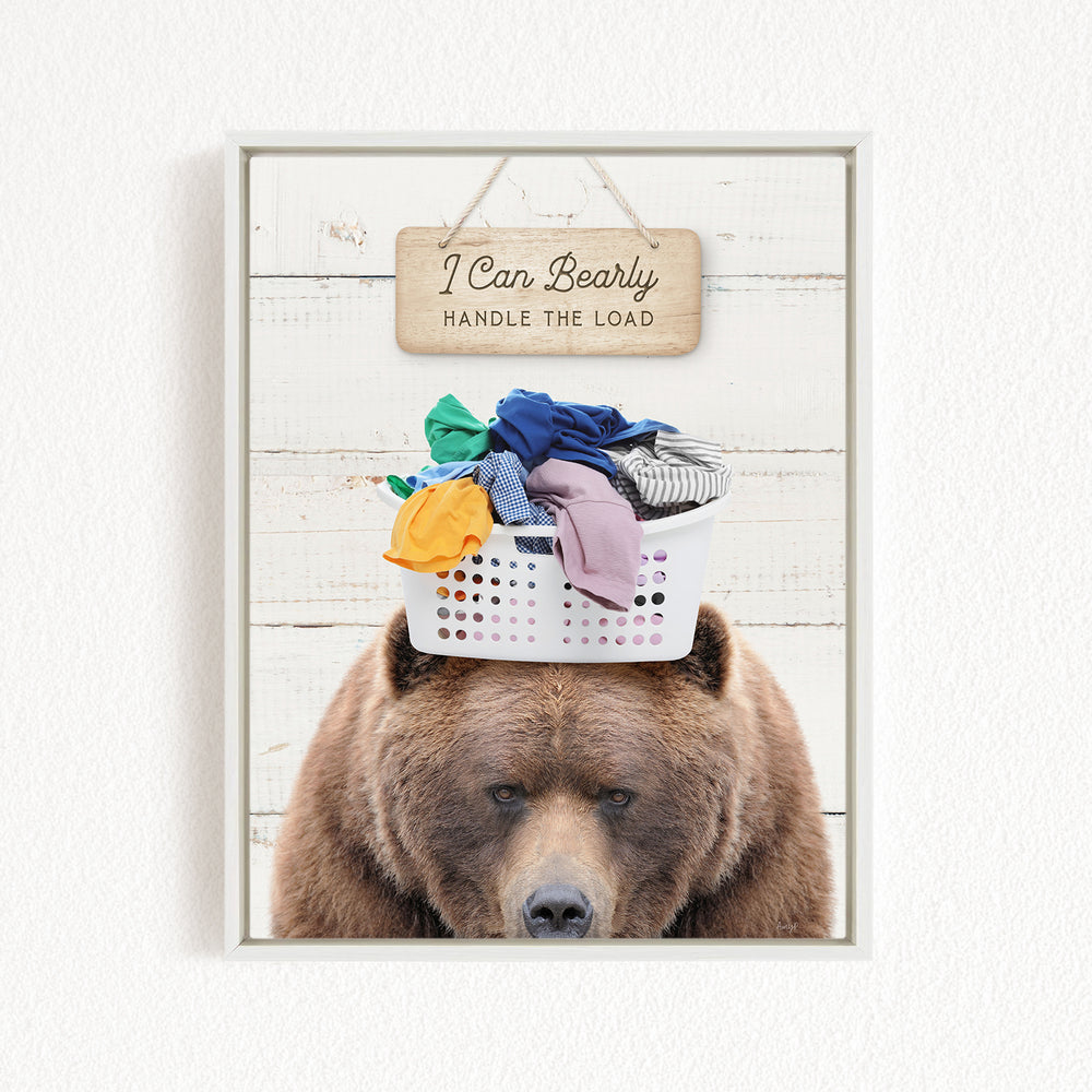 A brown bear sits in front of a white laundry basket filled with clothes, with a wooden sign above that reads "I Can Bearly Handle the Load".
