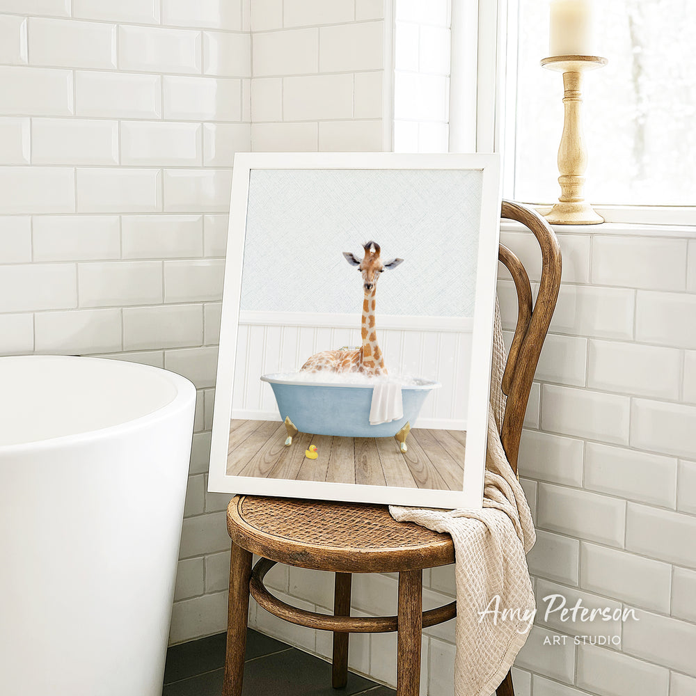 A framed image of a giraffe sitting in a bathtub, surrounded by white tiles, is displayed on a wooden chair in a bathroom setting.
