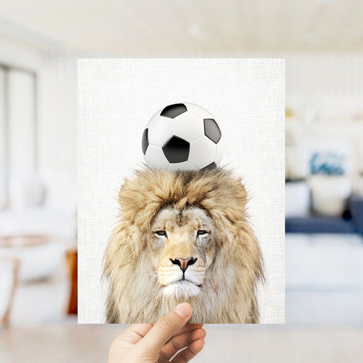 A lion with a soccer ball on its head is being held up in a room with a blurred background.
