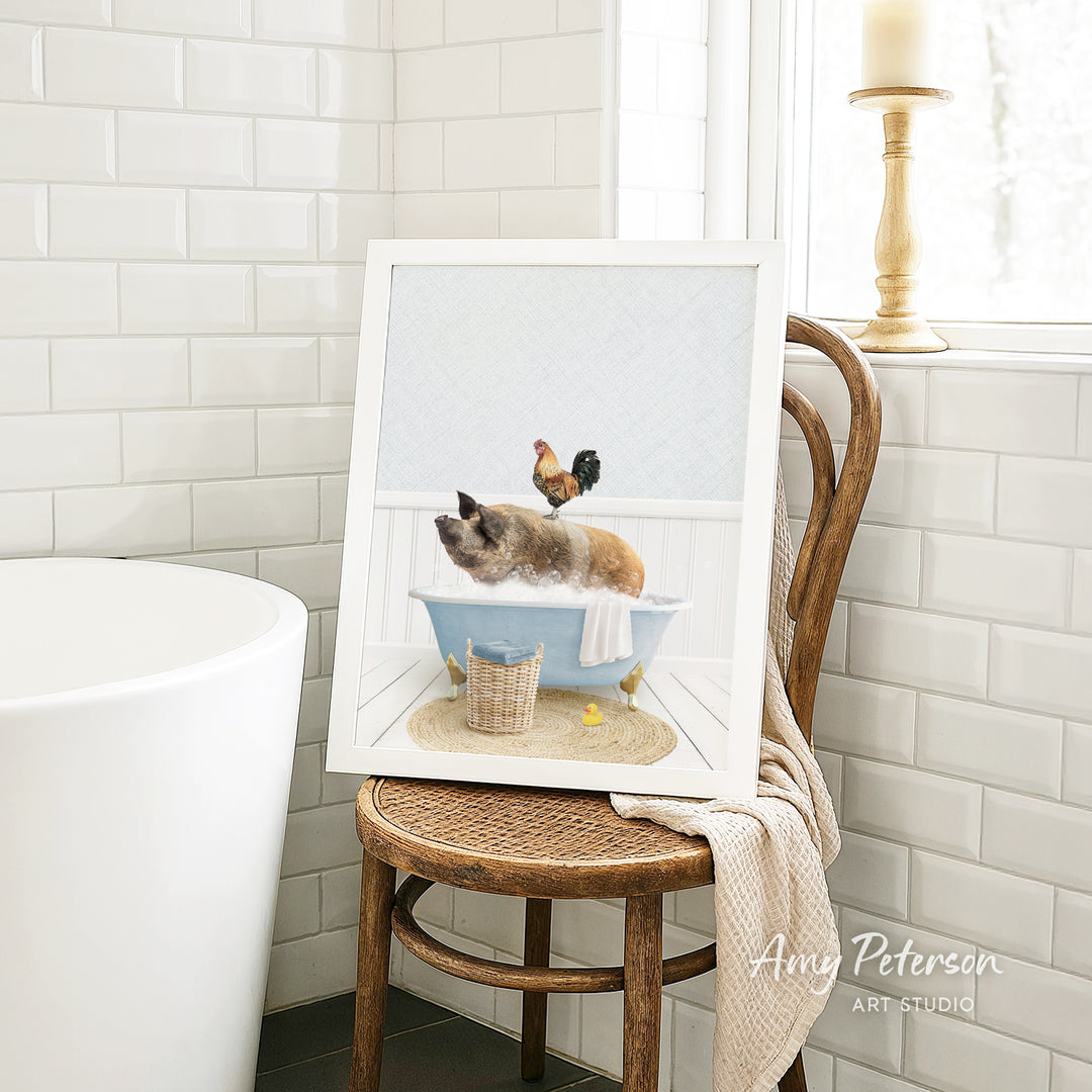 A framed image of a cat sitting in a bathtub, with a rooster perched on its back, is displayed on a wooden chair in a bathroom setting.