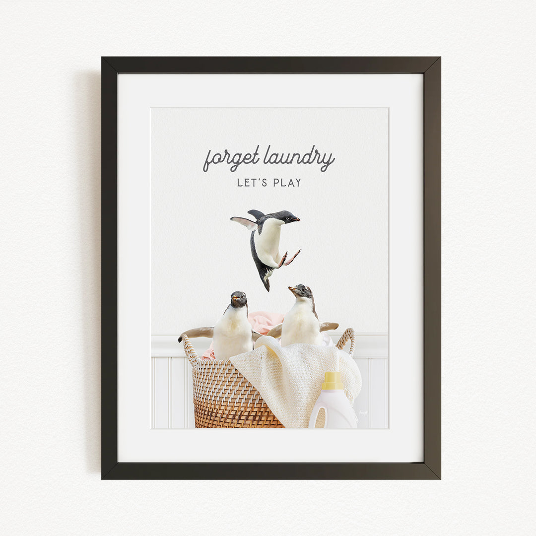 A framed image depicting three penguins in a basket, with the text "forget laundry, let's play" written above them.