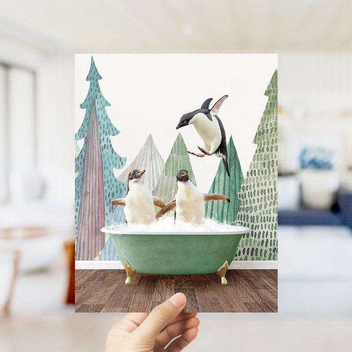 A hand holds up a framed image depicting two penguins sitting in a green bathtub, surrounded by a snowy landscape with trees and a bird in mid-flight.