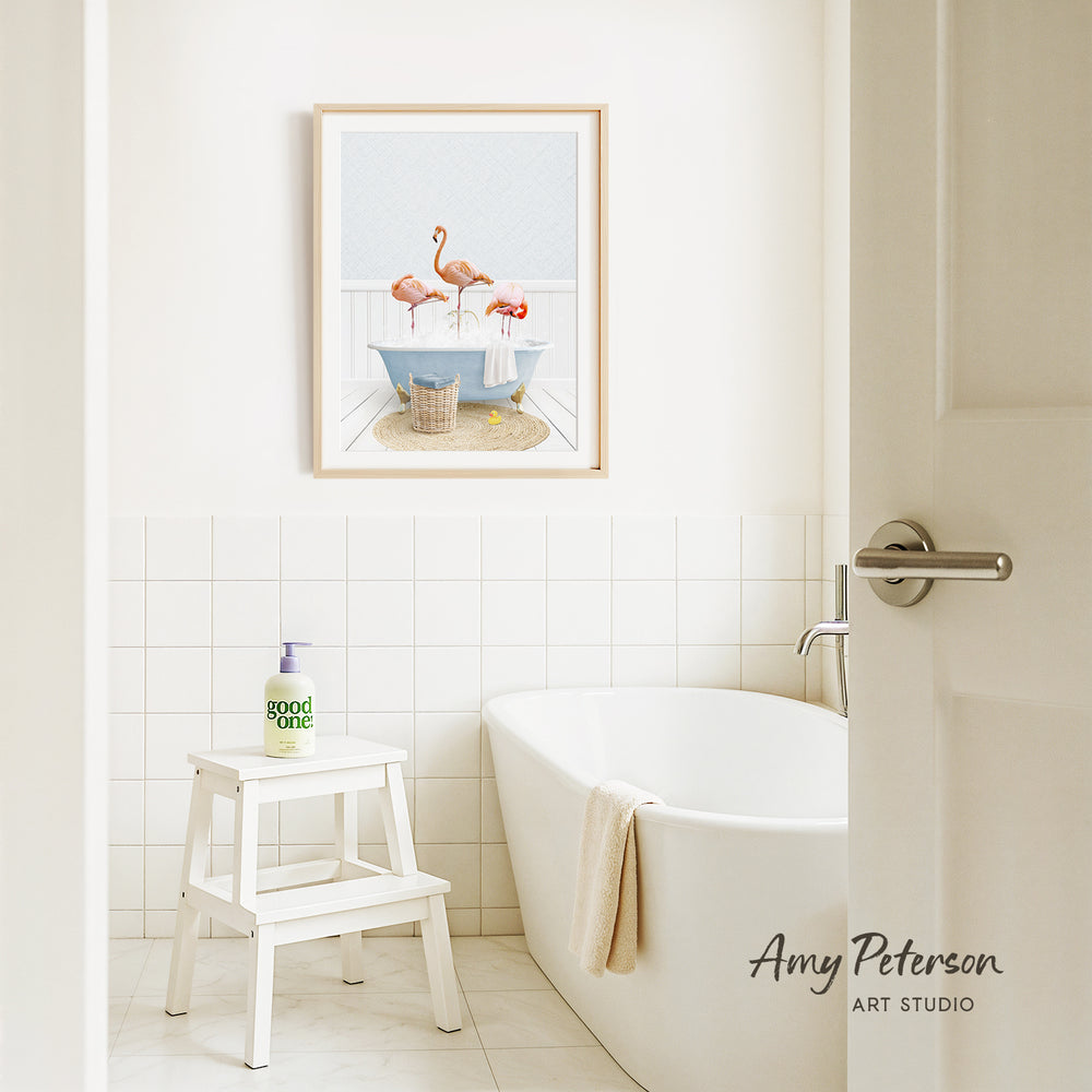 A bathroom with a white bathtub, a stool, and a framed picture of flamingos in a bathtub. The bathroom has white tiles and a white door.