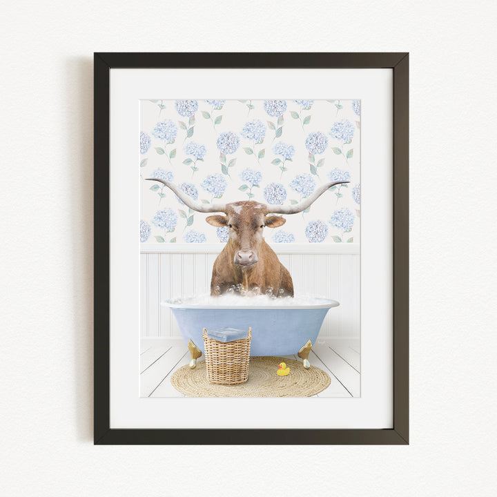 A brown cow with large horns is sitting in a blue bathtub filled with water, surrounded by a floral patterned wall and a basket of rubber ducks.