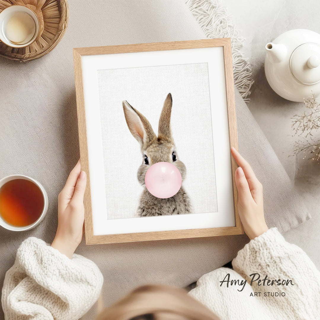 A person is holding a framed picture of a rabbit blowing a bubble with a pink bubblegum bubble, with a teapot and a cup of tea nearby.