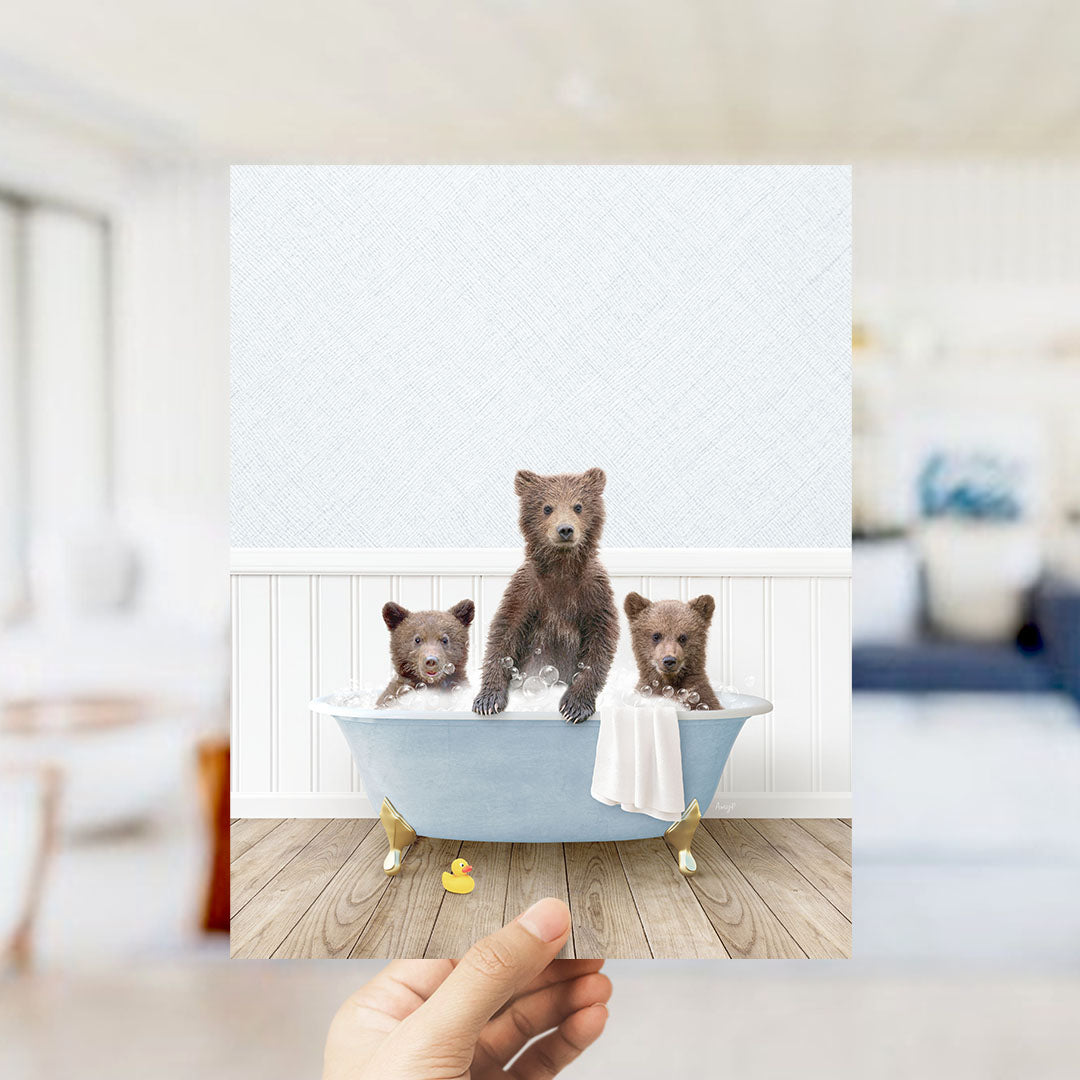 A hand holds up a framed image of three adorable brown bears sitting in a blue bathtub, surrounded by a white bathtub, a yellow rubber duck, and a white towel.