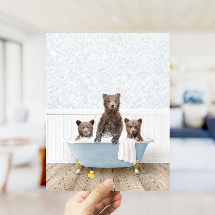 A hand holds up a framed image of three adorable brown bears sitting in a blue bathtub, surrounded by a white bathtub, a yellow rubber duck, and a white towel.