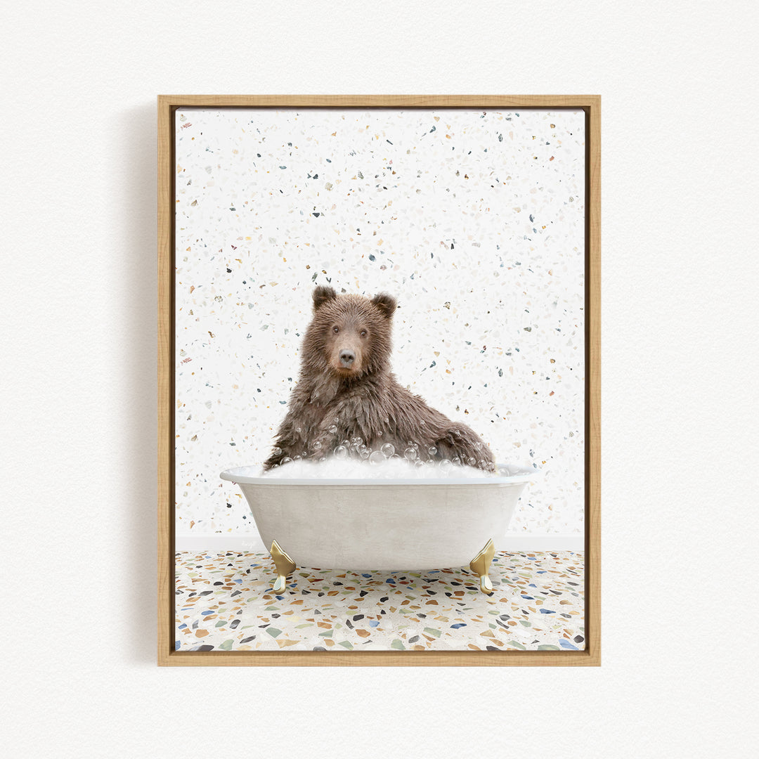 A brown bear sitting in a white bathtub filled with bubbles, surrounded by a speckled patterned floor.