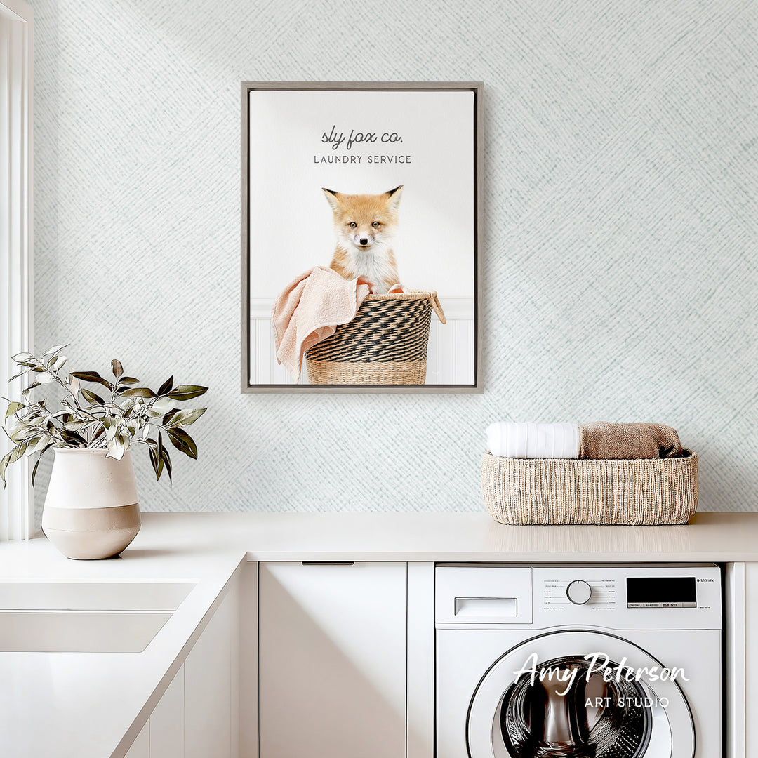 a framed photo of a cat sitting in a basket, with the text "sky fox co. Laundry service" above it. The photo is displayed on a wall above a white countertop with a potted plant and a basket of towels.