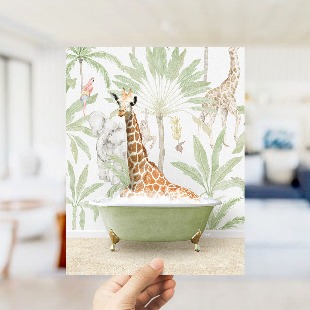 A hand holds up a framed print featuring a giraffe sitting in a bathtub surrounded by tropical foliage.