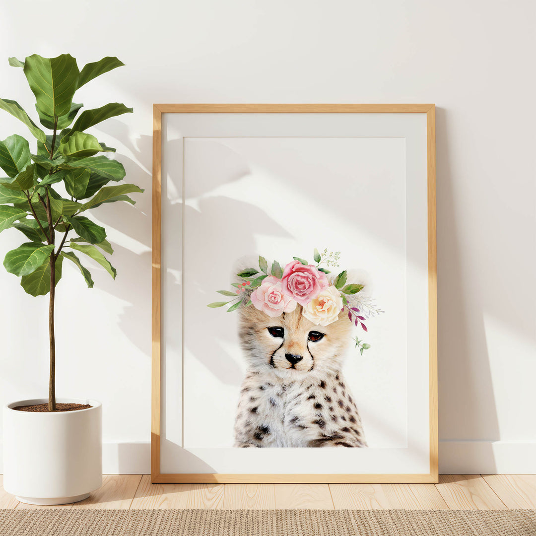 A framed print of a cheetah wearing a floral crown is displayed on a white wall, with a potted plant to the left.