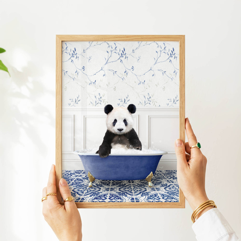A person's hands are holding a framed picture of a panda bear sitting in a blue bathtub, with a floral pattern on the wall behind it.