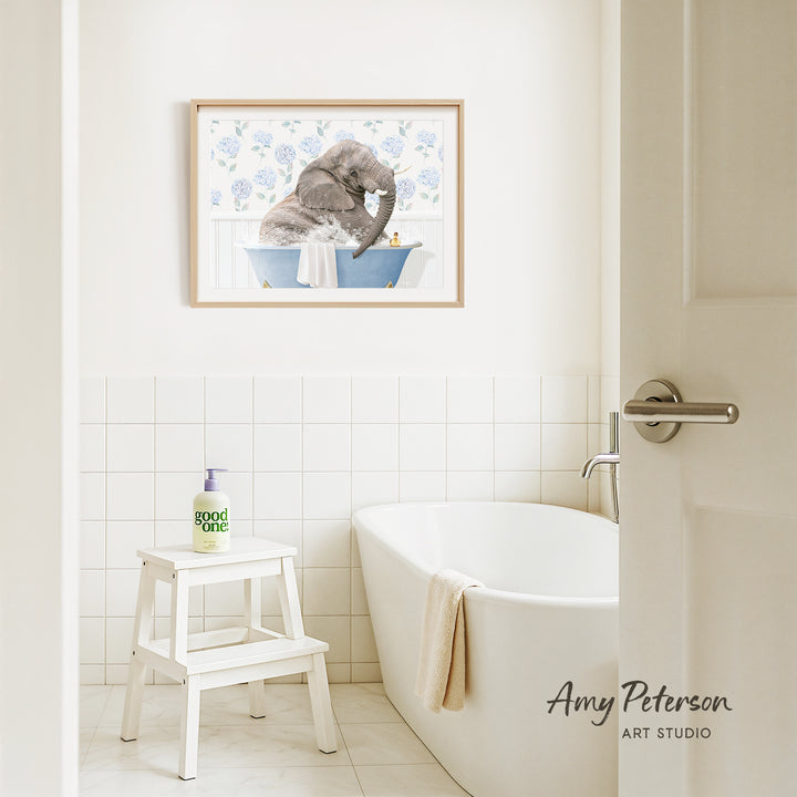 A framed painting of an elephant bathing in a tub hangs on a white tiled wall above a white bathtub in a bathroom.