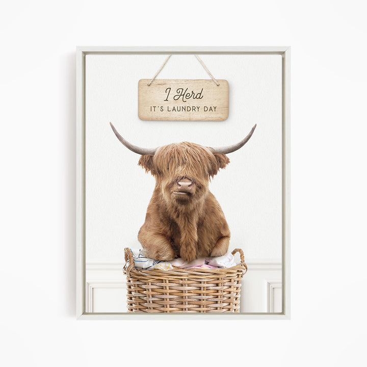 A brown Highland cow with shaggy fur sits in a wicker basket, with a wooden sign above it that says "It's laundry day".